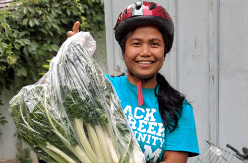 Mildred, staff at BCCF, holding some veggies in a bag and smiling. She is wearing a teal BCCF t-shit!