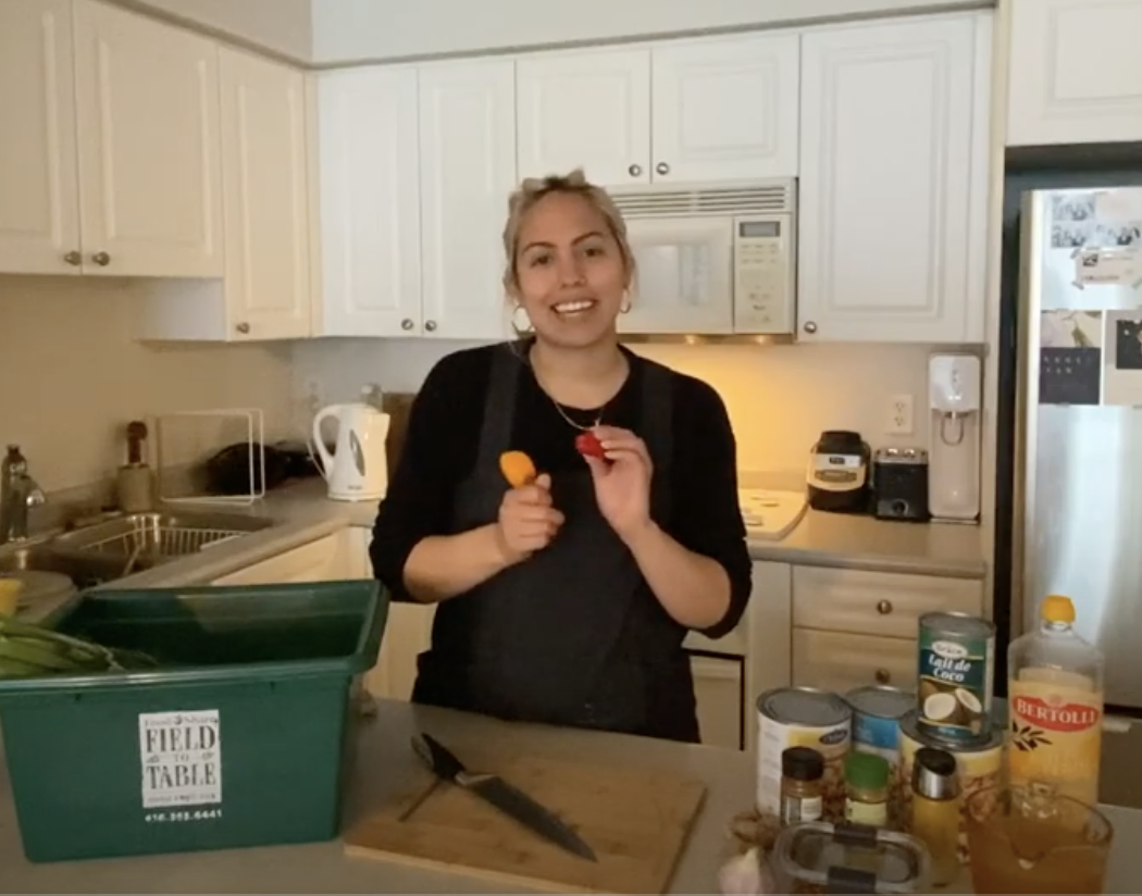 jade guthrie (woman) pictured, with a green food basket to her left, and many pre-cut vegetable to the right, ready to start cooking them. She holds in her hands an onion and a purple vegetable.