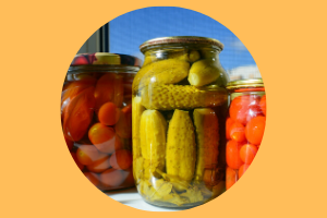 various pickles in glass jars, in a circular image with an orange background behind it.