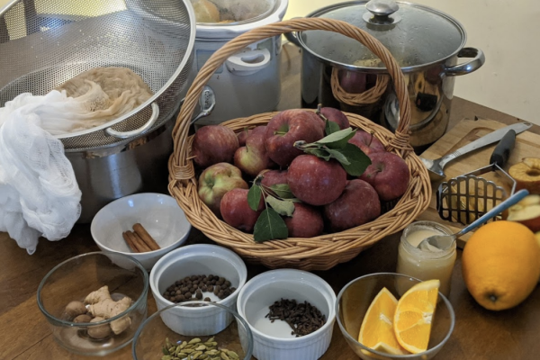 apples in a basket on a wooden table, surrounded by other ingredients in small white circular bowls: cinnamon, oranges, spices