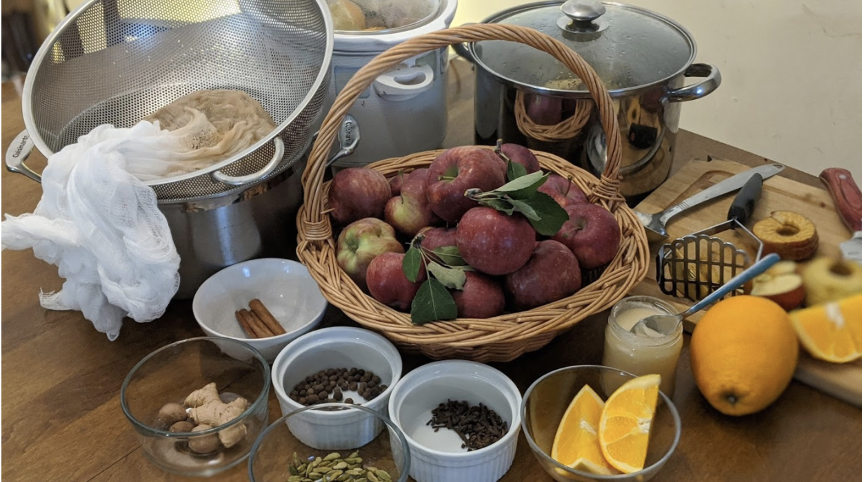 apples in a basket on a wooden table, surrounded by other ingredients in small white circular bowls: cinnamon, oranges, spices