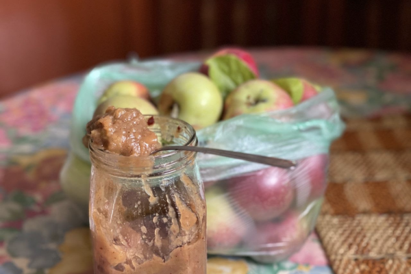 Jar of apple chutnety with sppon inside, bag of apples in hte background blurred out a bit, all istting on top of a wooden table