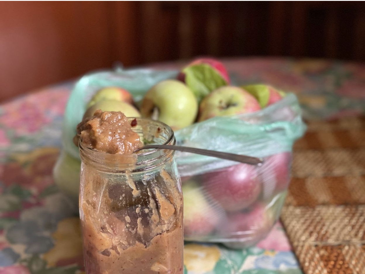 Jar of apple chutnety with sppon inside, bag of apples in hte background blurred out a bit, all istting on top of a wooden table